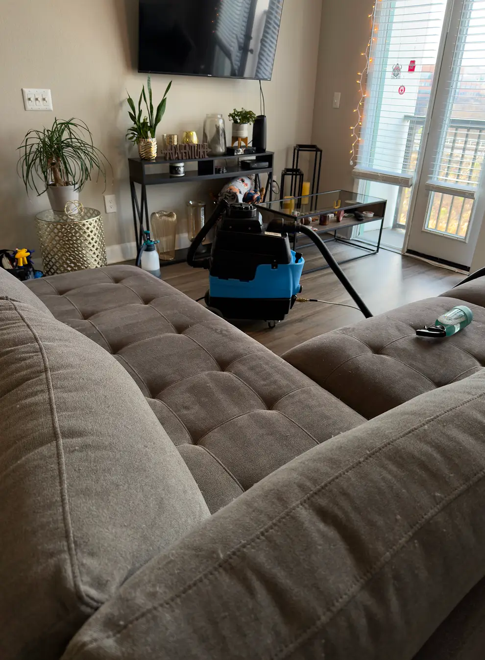 In-home upholstery cleaning process setup in a living room with a professional extraction machine and hoses near a large sectional couch, showing real couch cleaning service work and attention to detail.