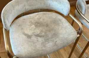 Cream accent chair before chair cleaning, with dark deep stains soaked into the seat and worn dull fabric.