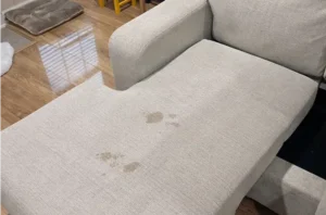 Light gray chaise before couch stain removal, showing a dried spill stain soaked into the seat fabric.